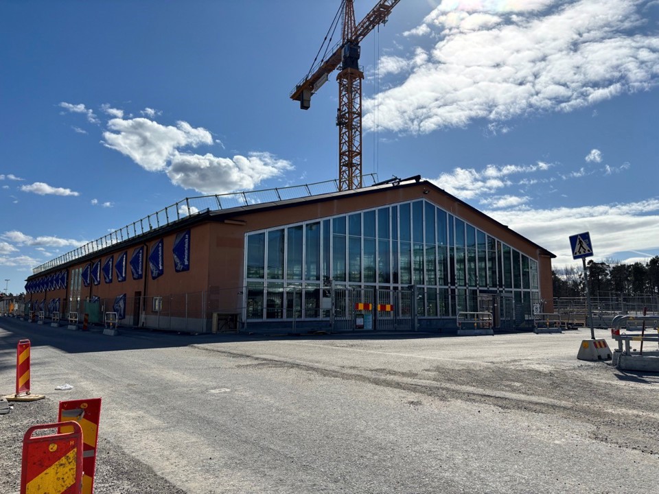 En byggarbetsplats med en orangefärgad byggnad under uppförande, med stora glaspartier. Blå himmel, med en byggkran i bakgrunden. Varnaringsmärken och skyddsbarriärer syns på vägen framför byggnaden.